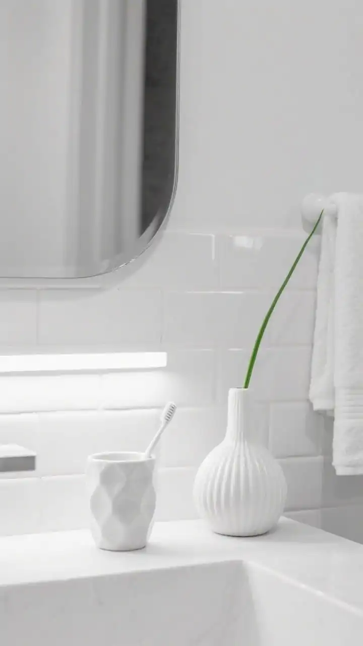 Detail closeup, straight-on macro composition for Modern Monochrome: white-on-white palette showcasing layered textures glossy subway tile stickers on the wall, satin-finish painted wall nearby, and matte white towel edge in frame; sculptural pill-shaped mirror reflecting a hint of space; ribbed ceramic toothbrush cup and a faceted white vase with a single green stem on a minimal vanity ledge; a linear vanity light casting crisp, even illumination; gallery-clean, high-contrast whites with subtle shadows for depth.