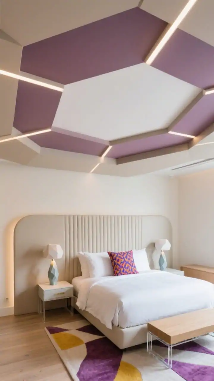 Overhead/oblique top-down angle highlighting a geometric layered ceiling of offset rectangular (or hexagon) panels at varied depths, color-blocked in soft taupe and dusty mauve with a central white panel; thin LED strips tracing select panel edges; below, a channel-tufted headboard; faceted ceramic lamps; slim modern furniture with powder-coated frames, light wood, and acrylic accents; graphic rug in complementary hues; bedding in solids with one bold patterned cushion; playful, polished boutique-hotel vibe.