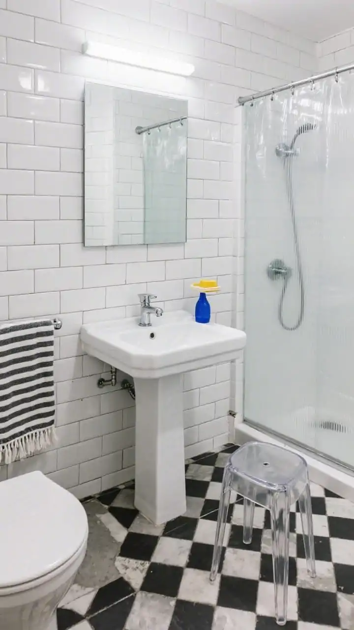Photorealistic wide overhead angle focusing on high-contrast surfaces in a small bathroom: walls wrapped in high-gloss white subway tile with tight grout lines, floor in bold black-and-white geometric cement tile. A slim pedestal sink (or narrow floating vanity) with chrome fixtures, a frameless mirror cabinet above, a clear shower curtain or glass panel with chrome rainfall plus handheld. Add a striped Turkish towel, a clear acrylic stool that visually disappears, and tiny pops of cobalt blue or sunny yellow via a soap dish or small vase. Bright, crisp linear LED lighting.