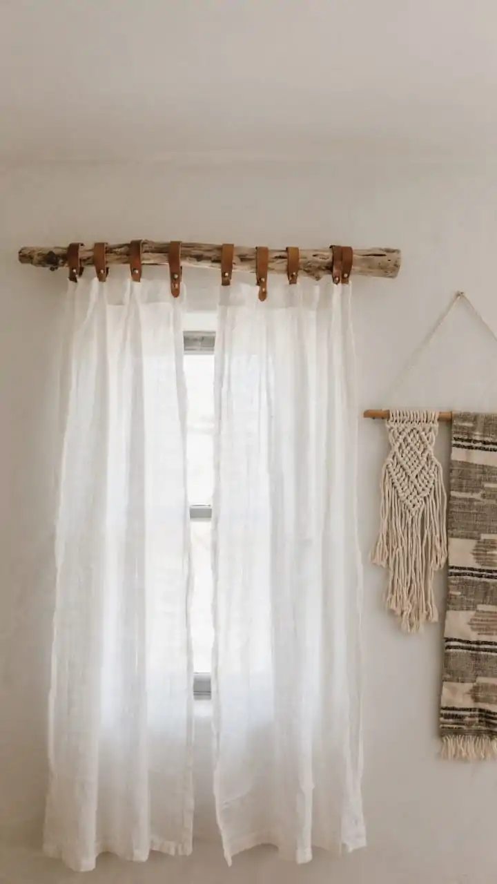 Medium, straight-on view of a wall display using a long piece of driftwood as a rod for textiles, styled like a boutique hotel. Lightweight white linen curtain panels hang at a window on the driftwood rod supported by leather straps with subtle patina; to the side, a woven macramé wall hanging and a vintage textile drape share the rod ends. Natural light filtering through gauzy fabric, neutral palette of whites, sand, and warm gray; materials include linen, cotton, leather, and raw wood; eco-chic, minimal styling without clutter.