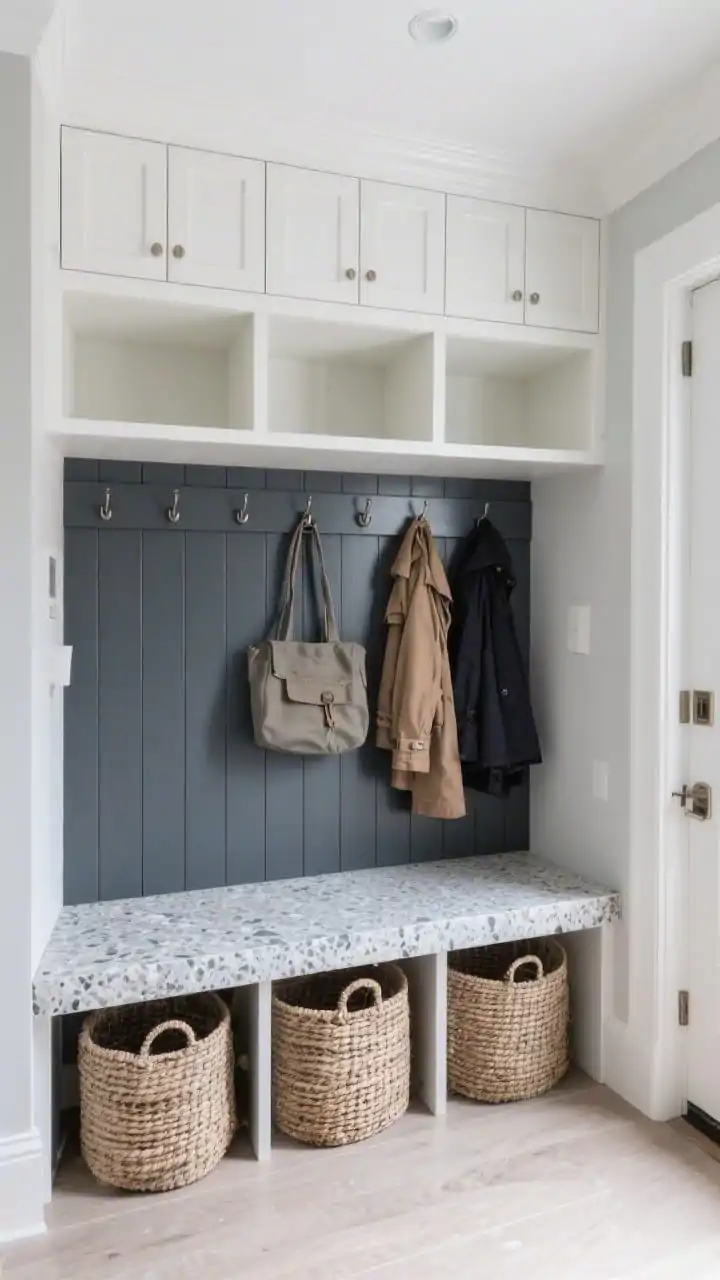 Wide shot, straight-on view of a polished mudroom-in-a-box entry: a custom built-in cubby wall with closed upper cabinets, open middle cubbies with integrated hooks holding bags and coats, and textured baskets below. A monolithic quartz or terrazzo bench forms a durable stone seat. The back panel is beadboard painted deep slate, framing crisp white and soft gray elements. Even, bright lighting highlighting the stone and beadboard textures. Photorealistic.
