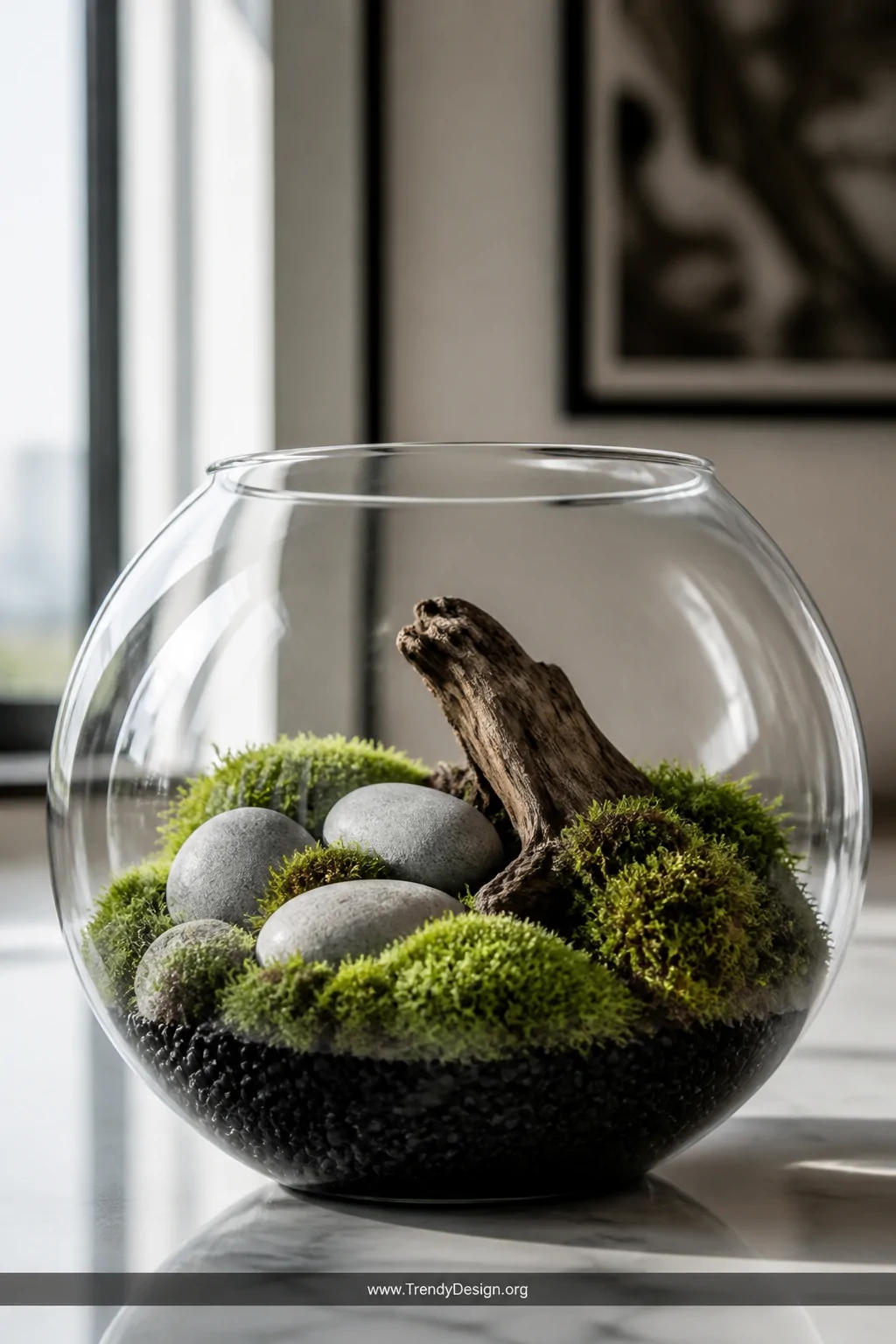 Close-up detail shot of a spherical glass fish bowl terrarium on a white marble surface, layered with glossy black pebbles at