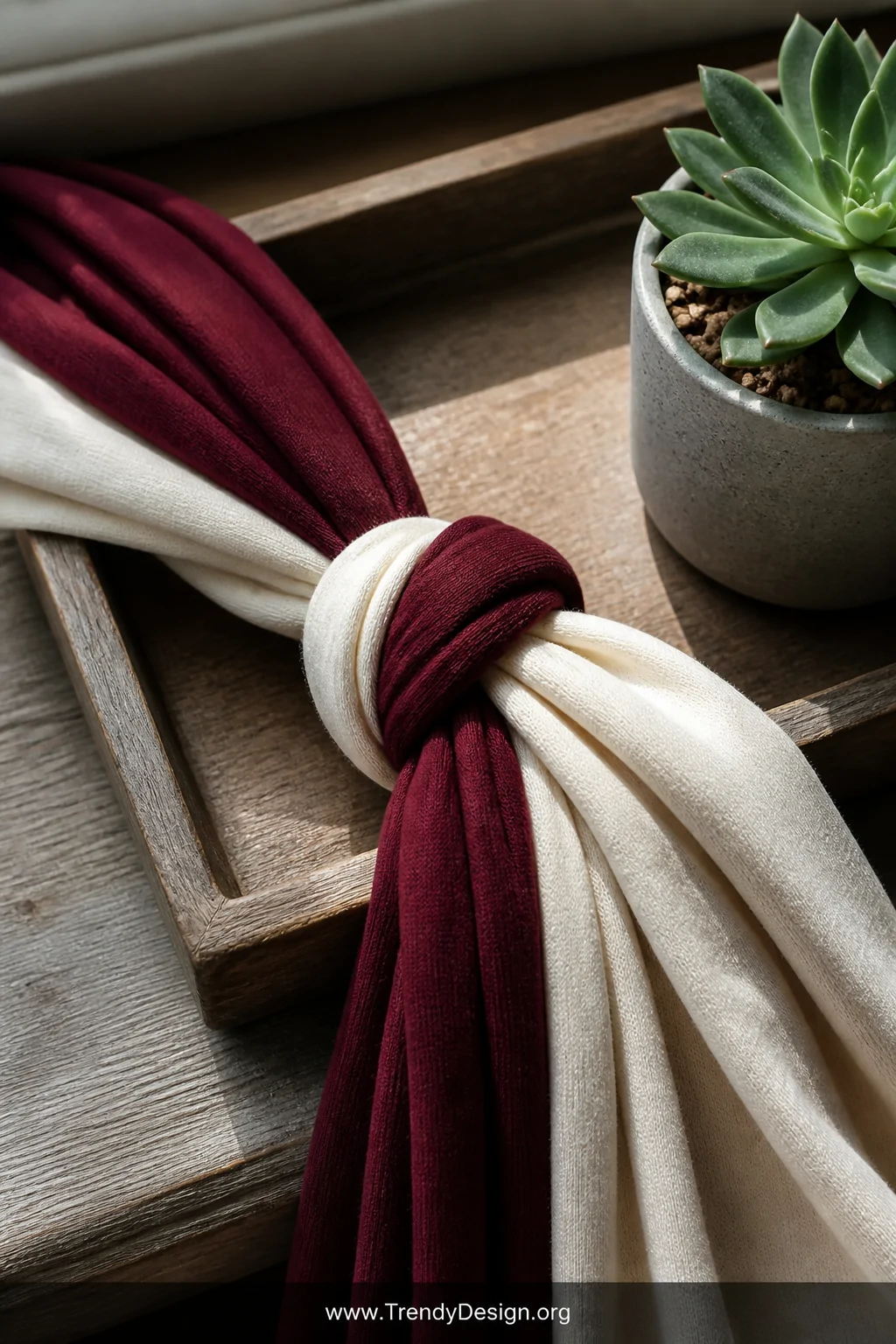 A closeup detail shot of two twisted jersey fabric strips in deep burgundy and cream, knotted at the center and draped over a