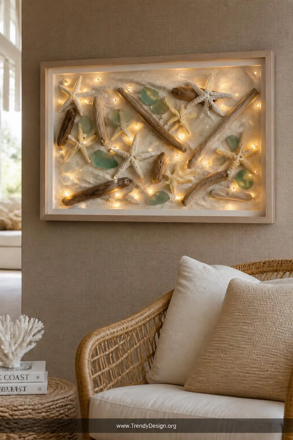 A wide shot of a coastal living room gallery wall centered on a deep shadowbox frame filled with white sand, arranged driftwo