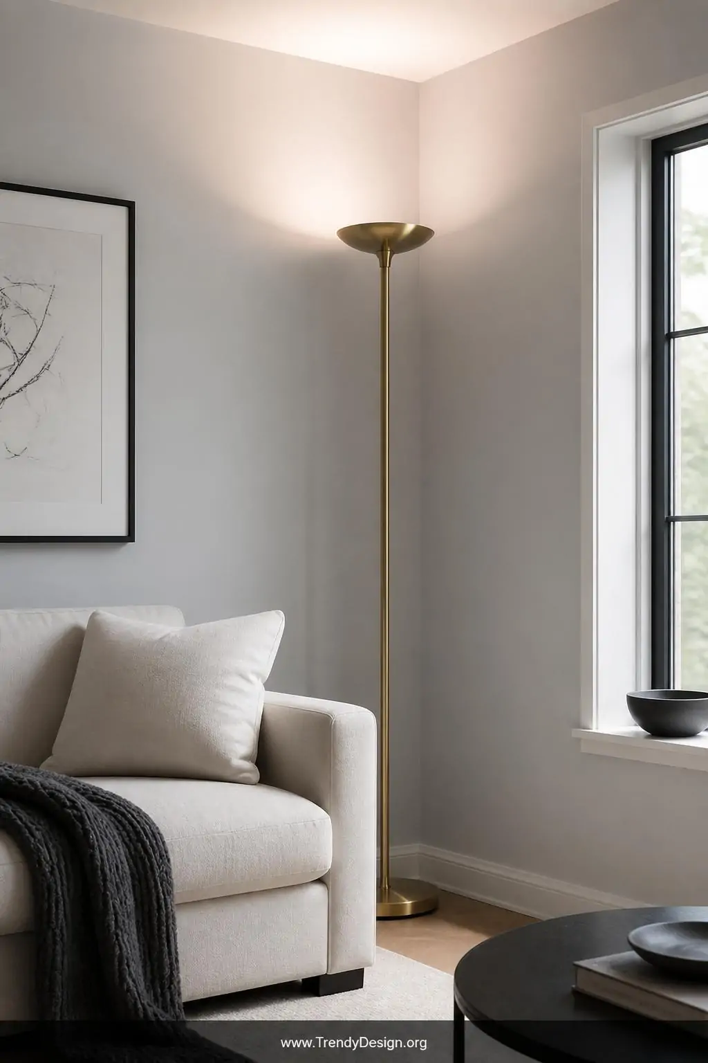 A slim brushed brass torchiere floor lamp stands in a tight apartment corner beside a minimalist linen sofa, directing soft d