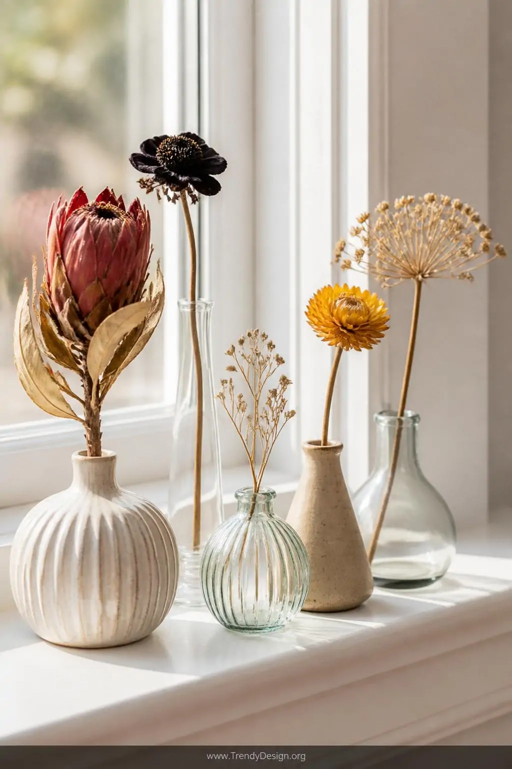 A tight closeup of five mismatched glass and ceramic bud vases clustered on a sun-washed white windowsill, each holding a sin