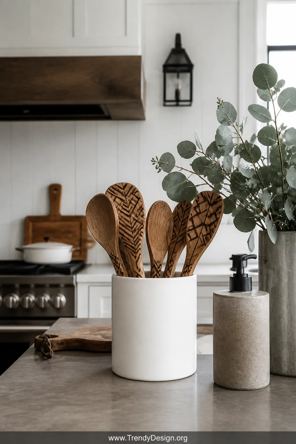 A modern farmhouse kitchen counter wide shot featuring a matte white ceramic utensil holder filled with wooden spoons burned 