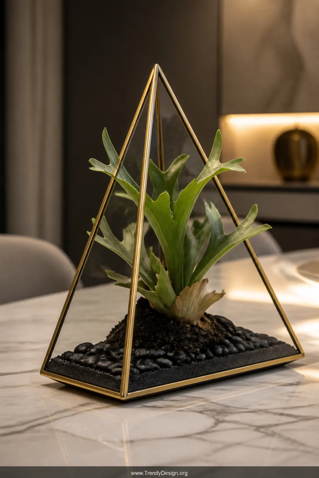Detail shot of a brass-framed geometric triangular prism terrarium sitting on a marble dining table, a single dramatic stagho