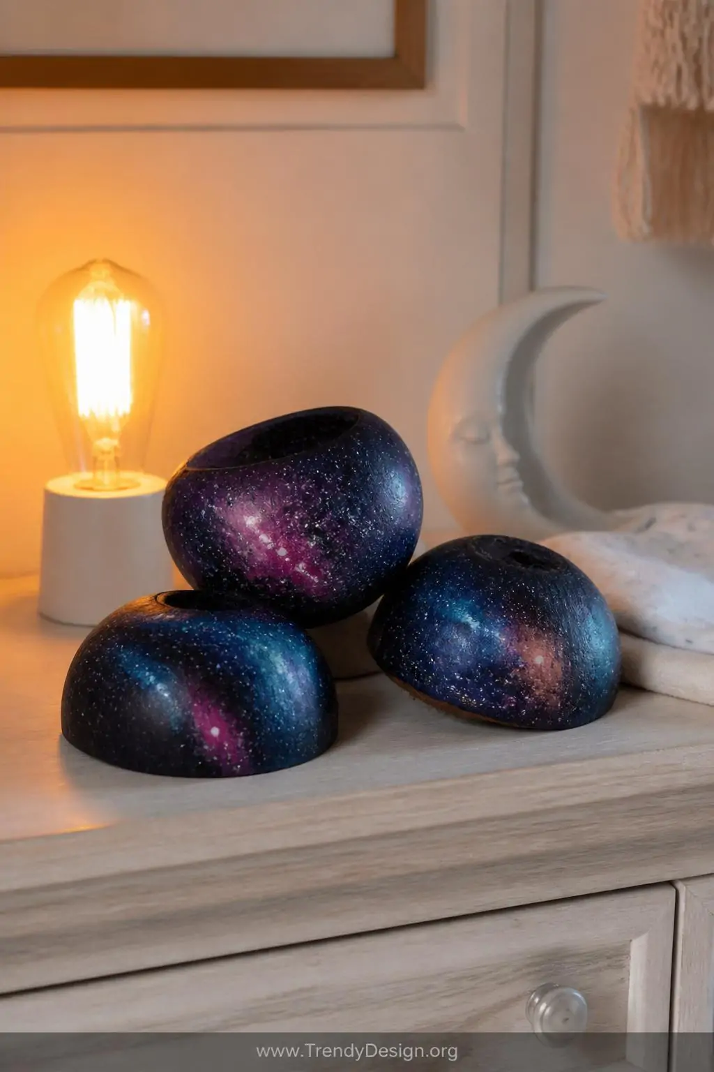 Close-up shot of three galaxy-painted coconut shells resting on a white oak nursery dresser, their deep navy and black surfac