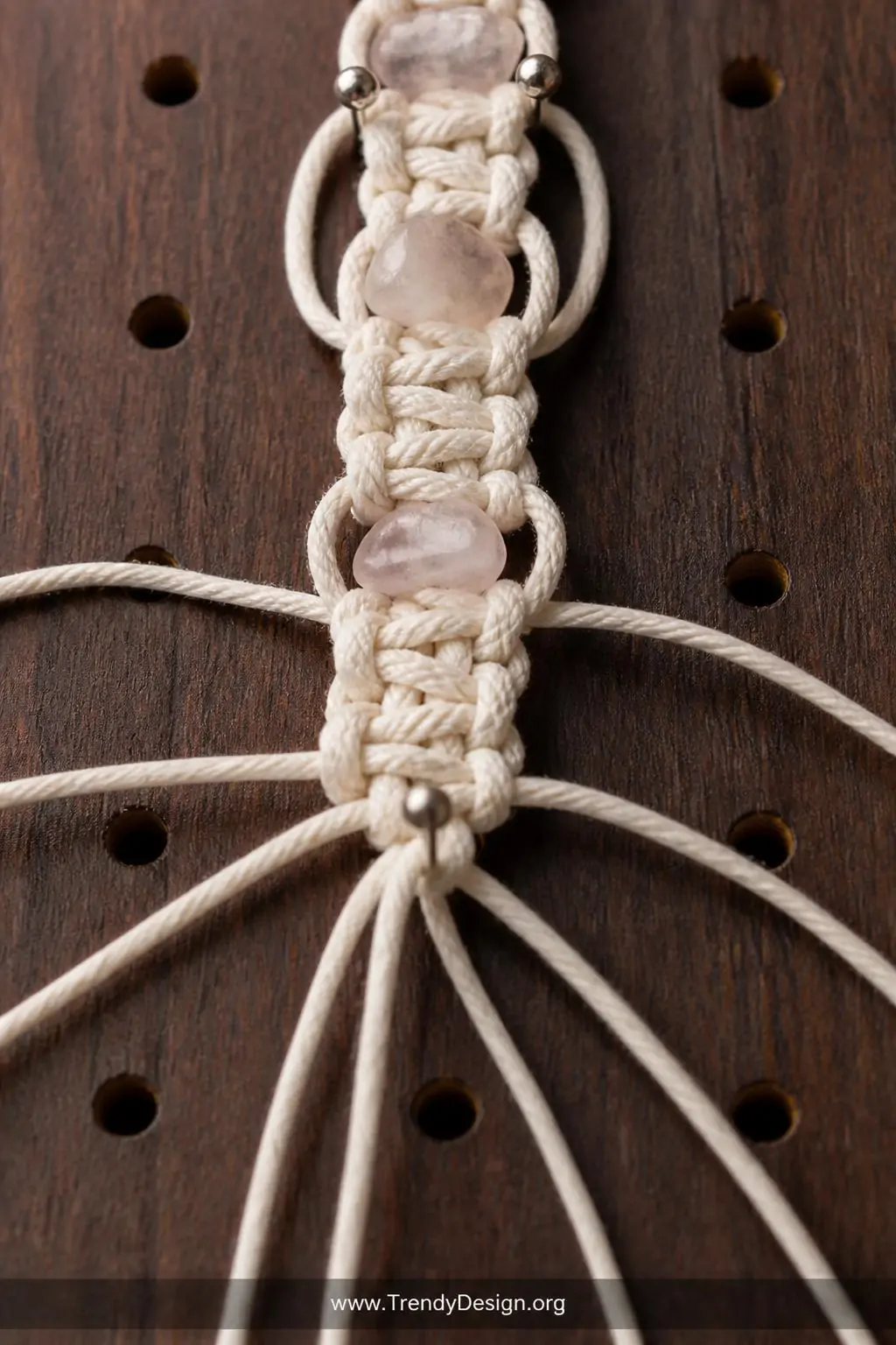 A closeup shot reveals the intricate knotwork of a natural cotton macramé bracelet mid-construction on a dark walnut pinboard