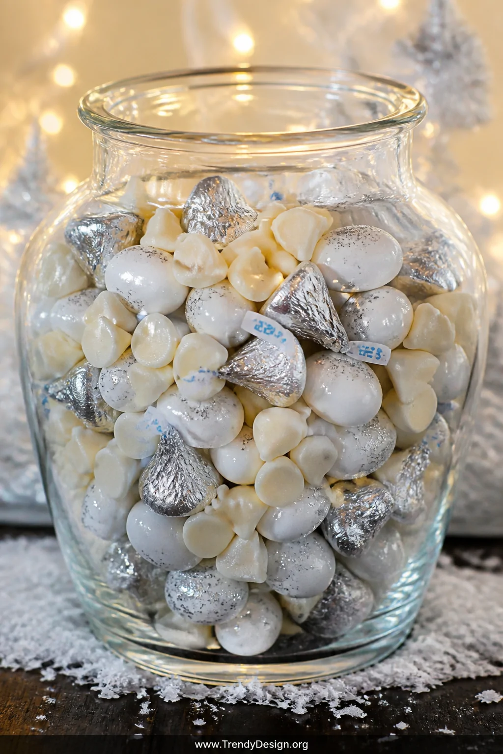 A detail closeup of a wide glass jar filled with white chocolate chips, silver-wrapped Hershey kisses, and white Jordan almon