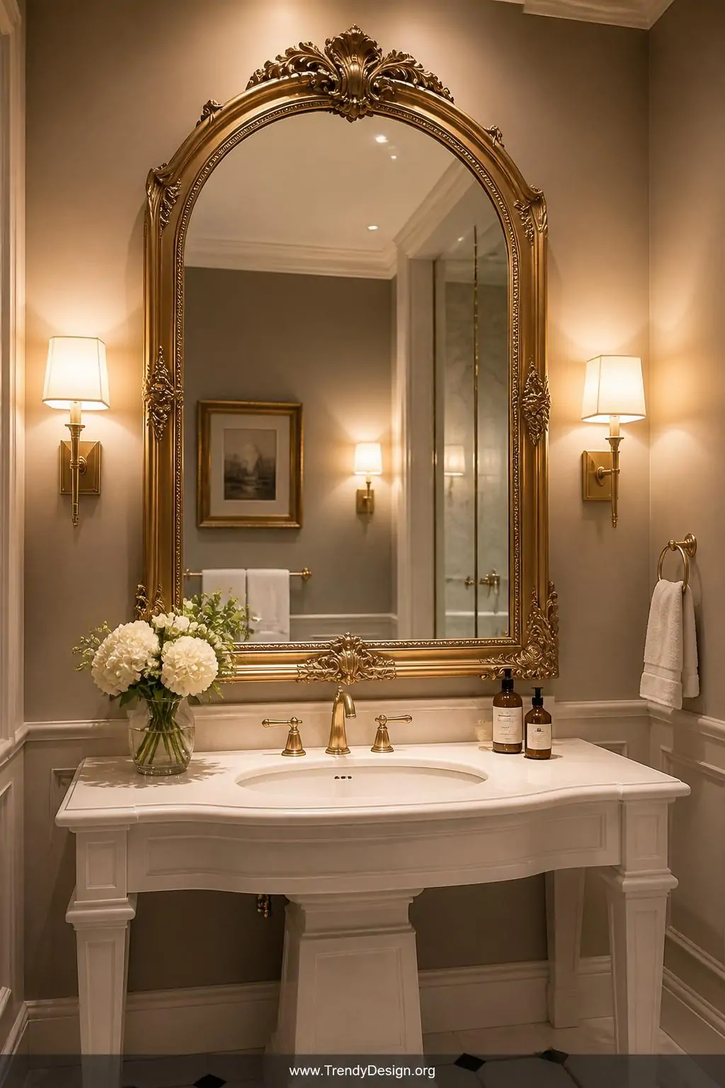 A dramatic arched ornate mirror with an antique gold frame mounted above a white pedestal vanity, medium shot showing how the