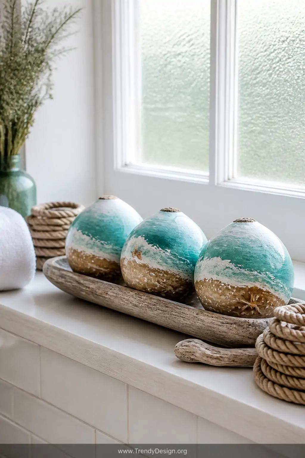 Medium shot of a breezy coastal bathroom windowsill displaying nautical coconut shells painted with turquoise and seafoam oce