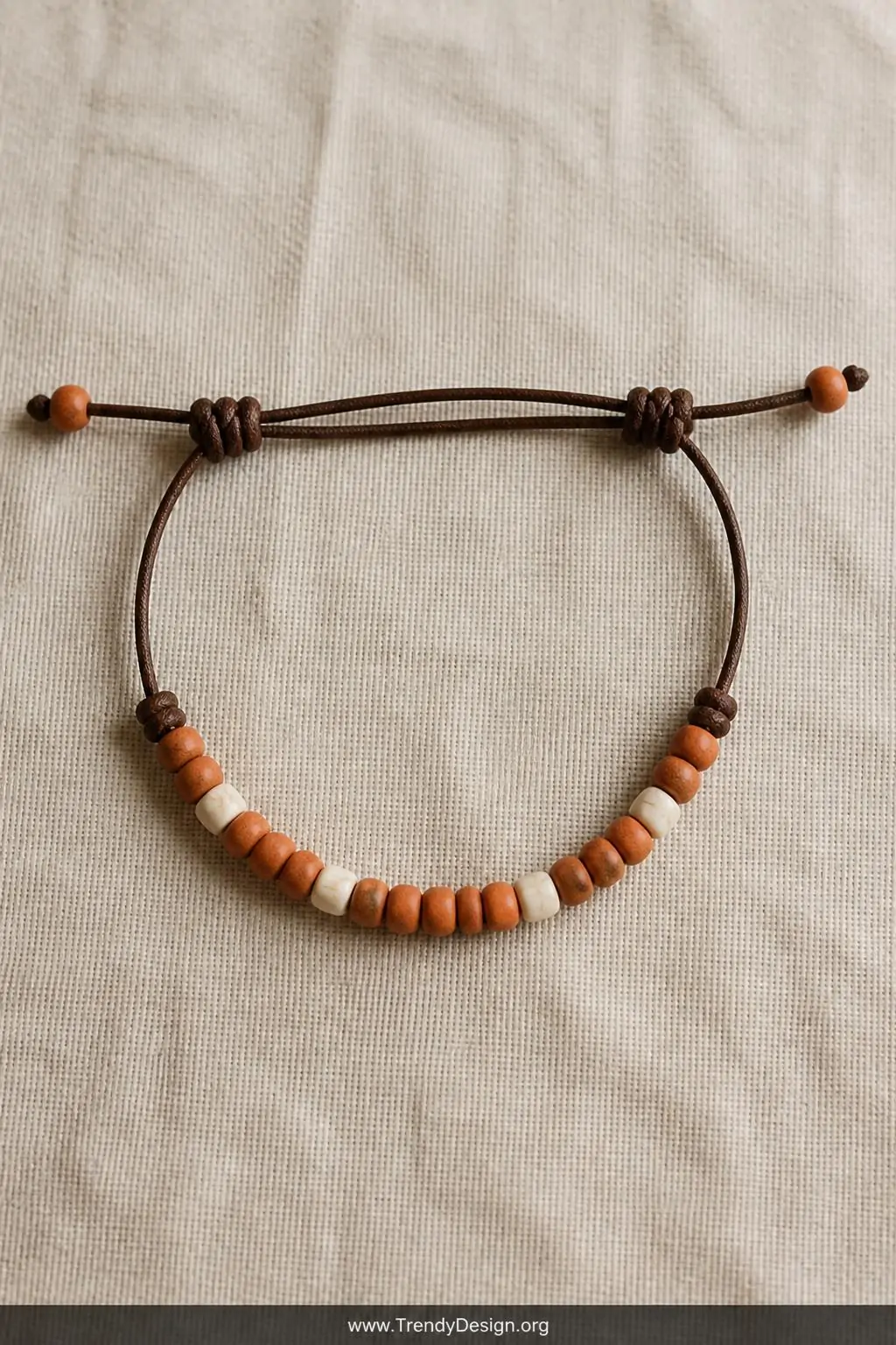 A natural linen-covered flat lay surface presents a leather cord bracelet with earthy terracotta and bone beads, two sliding 