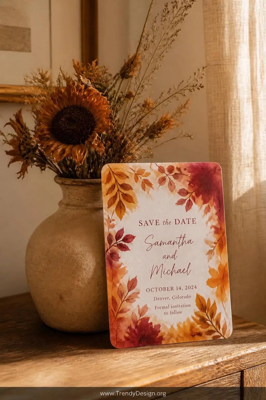 A medium shot of a seasonal watercolor save the date magnet propped against a small clay vase holding loose dried sunflowers,