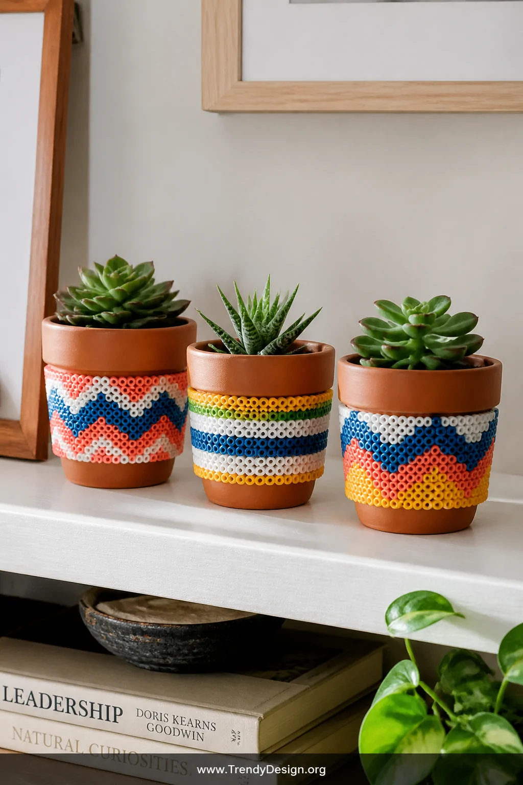 A styled shelf vignette close-up featuring small terra cotta plant pots wrapped in handmade Perler bead band borders in bold 