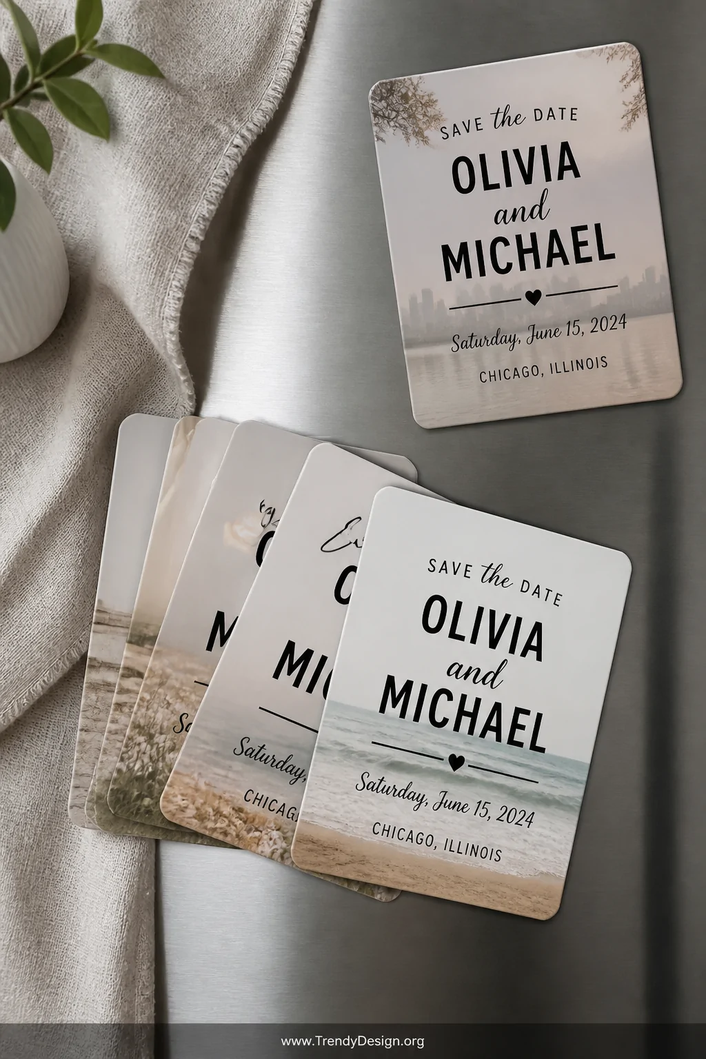 A wide flat-lay on a cool grey linen cloth displays a set of polished magnetic save-the-date cards printed with soft neutral 