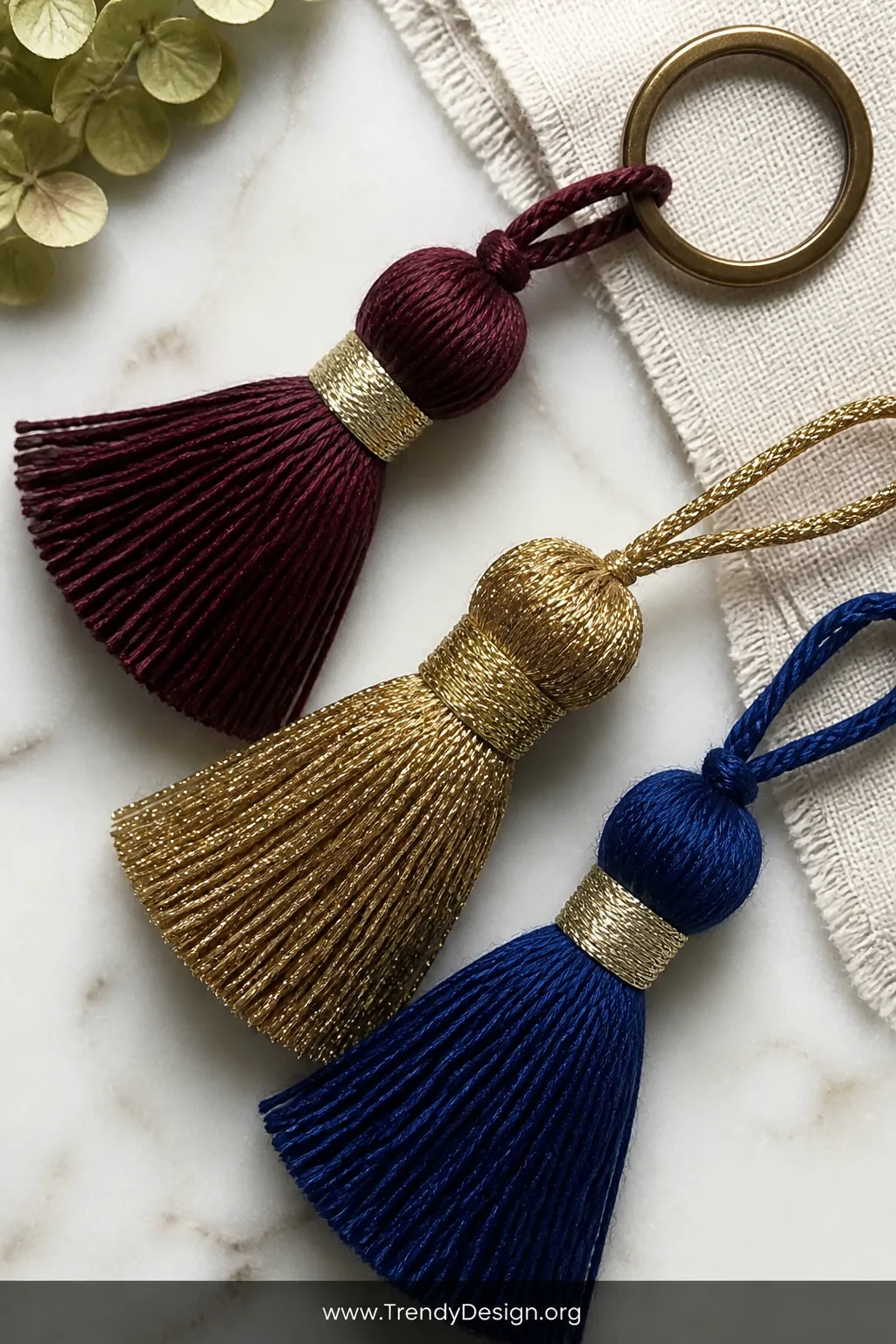 A flat lay close-up of handmade yarn tassel keychains in deep burgundy, gold metallic thread, and cobalt blue cotton arranged