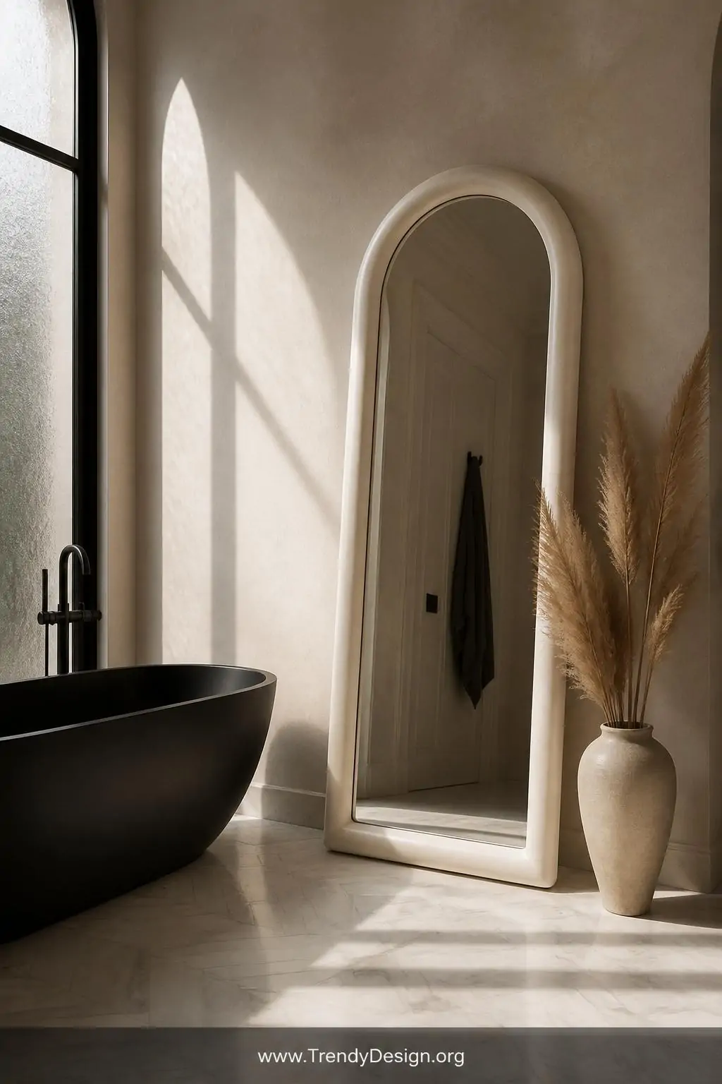 A tall slim arched mirror with a chunky plaster-white frame leaning beside a matte black freestanding soaking tub on pale her