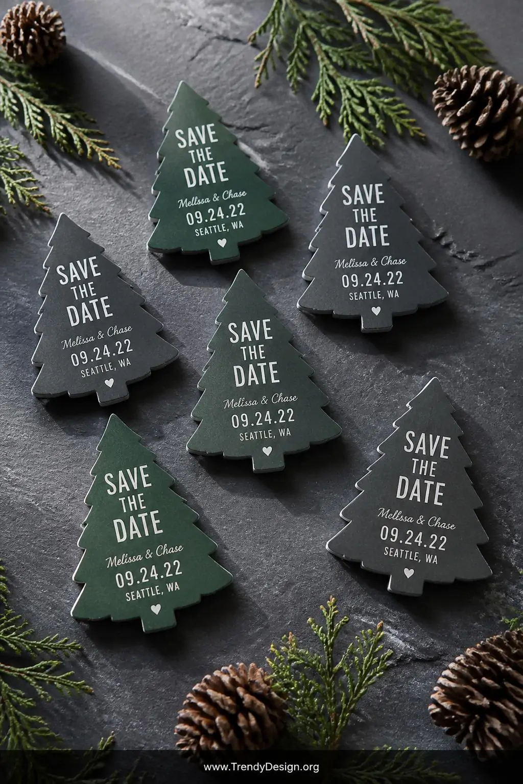 A wide shot of multiple pine tree silhouette die-cut save the date magnets arranged in a loose cluster on a raw slate surface