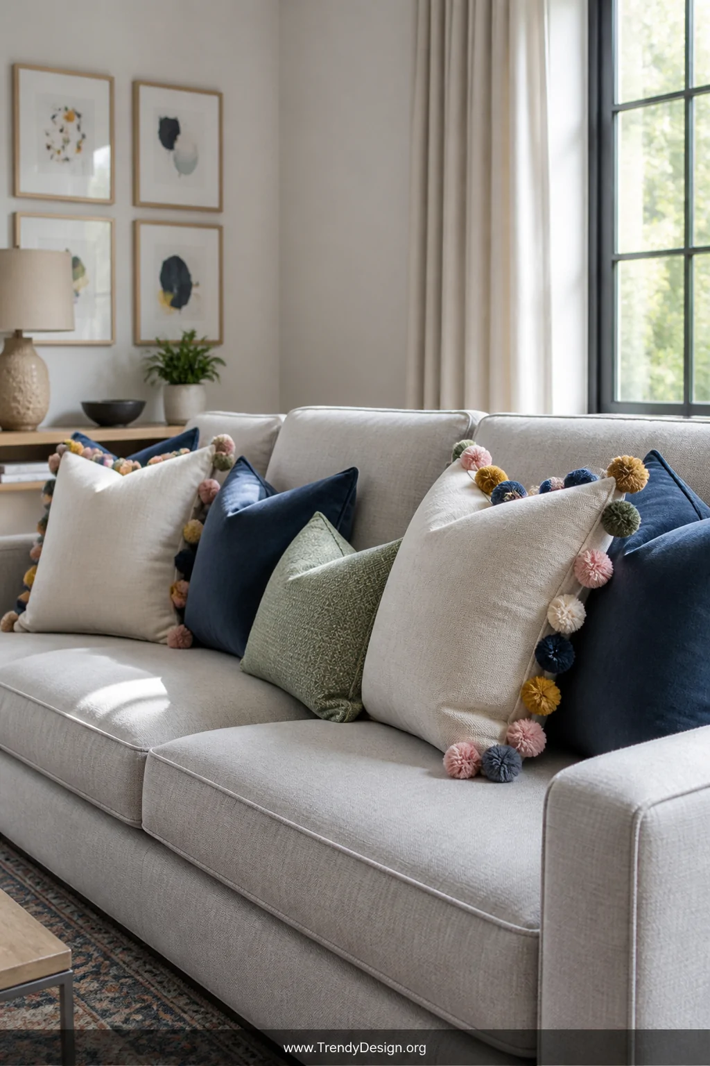 A medium shot of a dove gray linen sofa styled with a mix of plain pillow covers edged in multicolor oversized pom-pom trim a