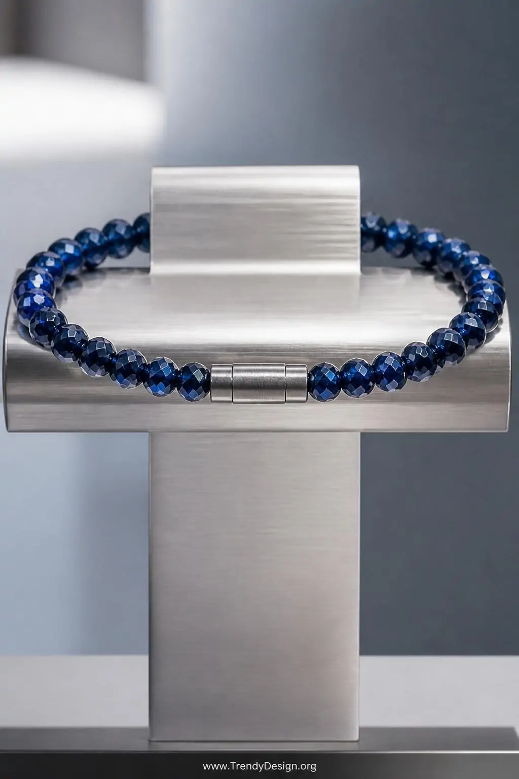 A brushed silver jewelry stand holds a single-strand deep sapphire crystal beaded bracelet where a cylindrical barrel clasp b