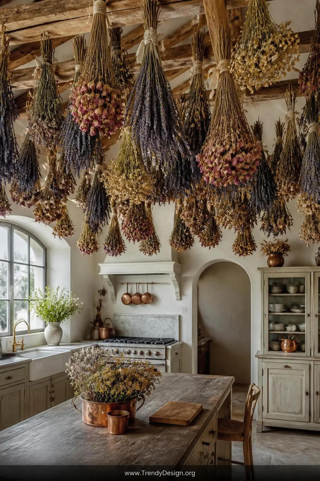A wide shot of a charming French-country kitchen with exposed ceiling beams from which hang multiple clustered bundles of ups