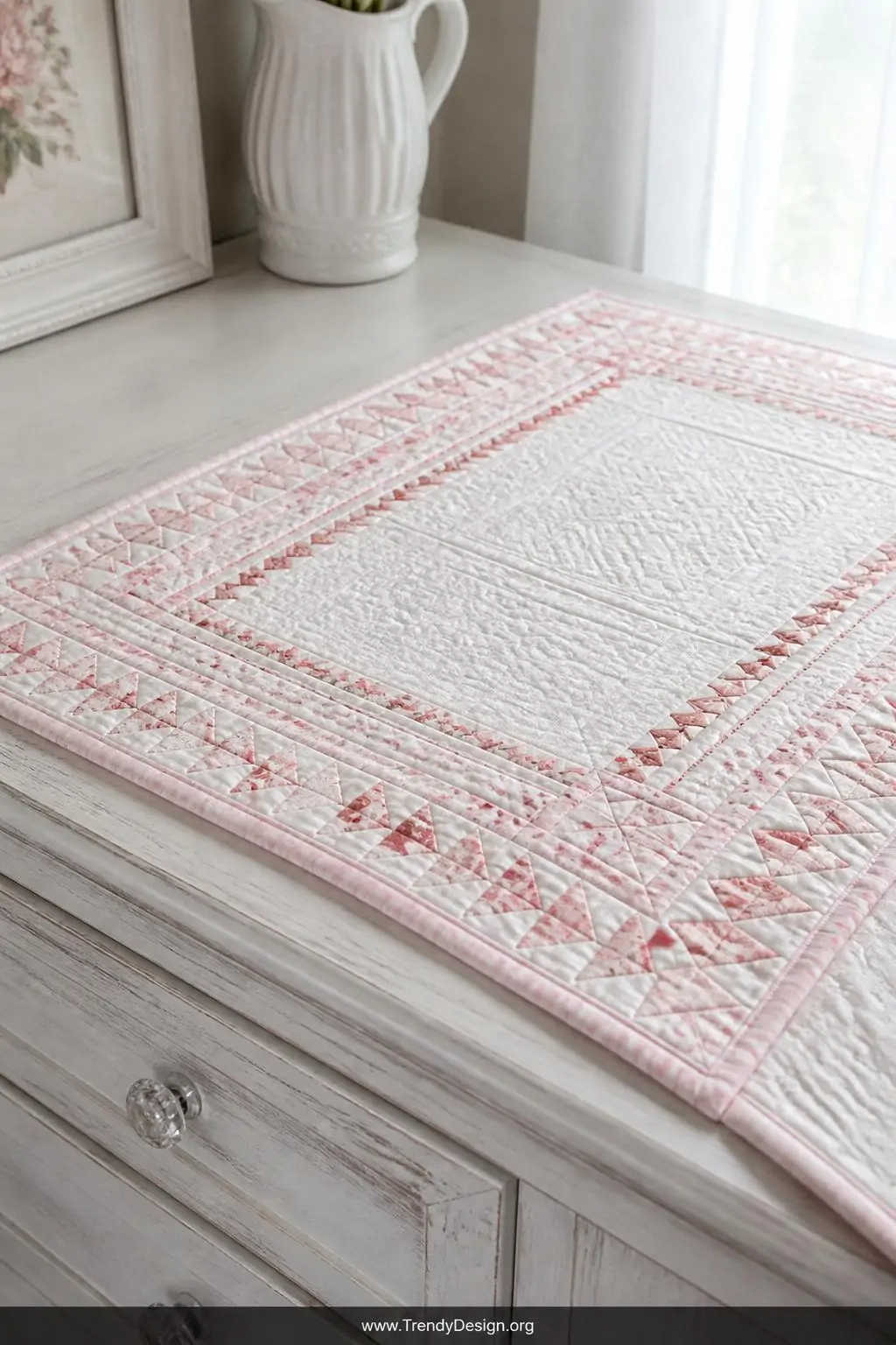 Closeup shot of a delicate baby quilt laid on a whitewashed nursery dresser surface, miniature flying geese border with indiv