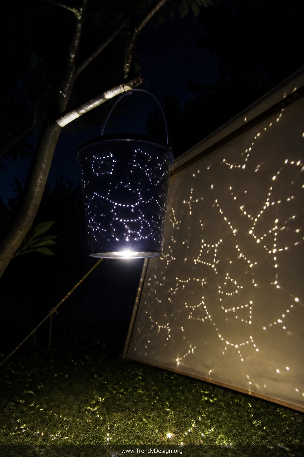 A dramatic medium shot of a dark navy blue bucket hanging from a tree branch at night, with dozens of tiny drilled constellat