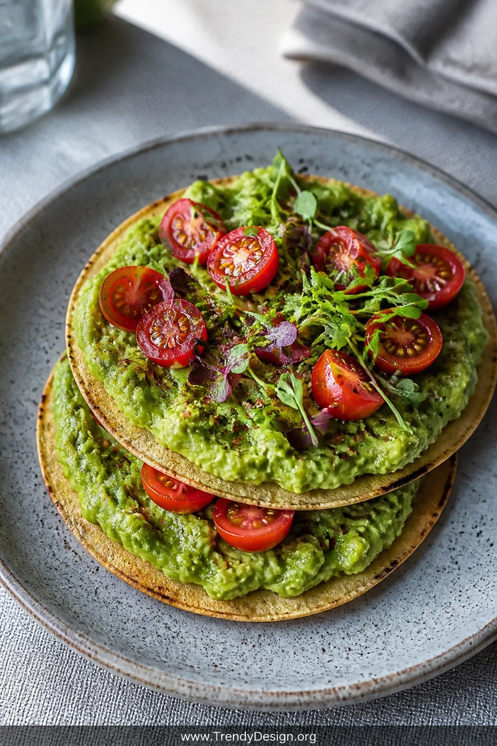 A close-up beauty shot of two stacked toasted corn tortillas on a speckled ceramic plate, generously topped with vivid green 