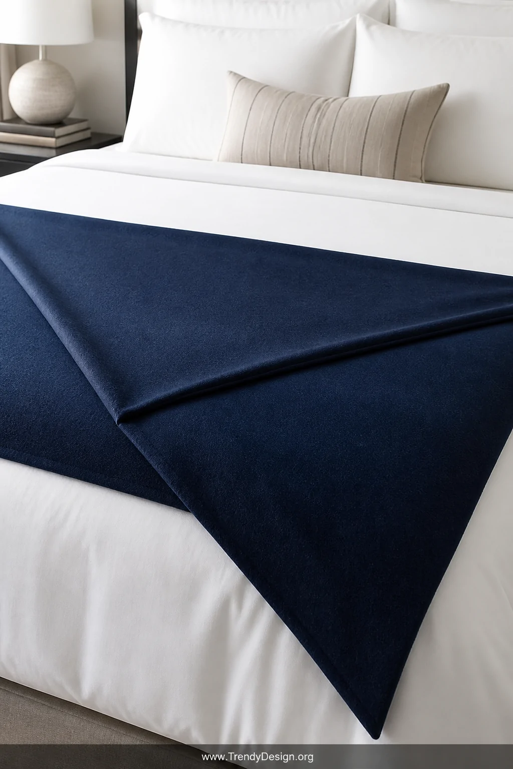 A close-up detail shot of a neatly made bed with crisp white linen bedding where a solid navy blue blanket has been folded in