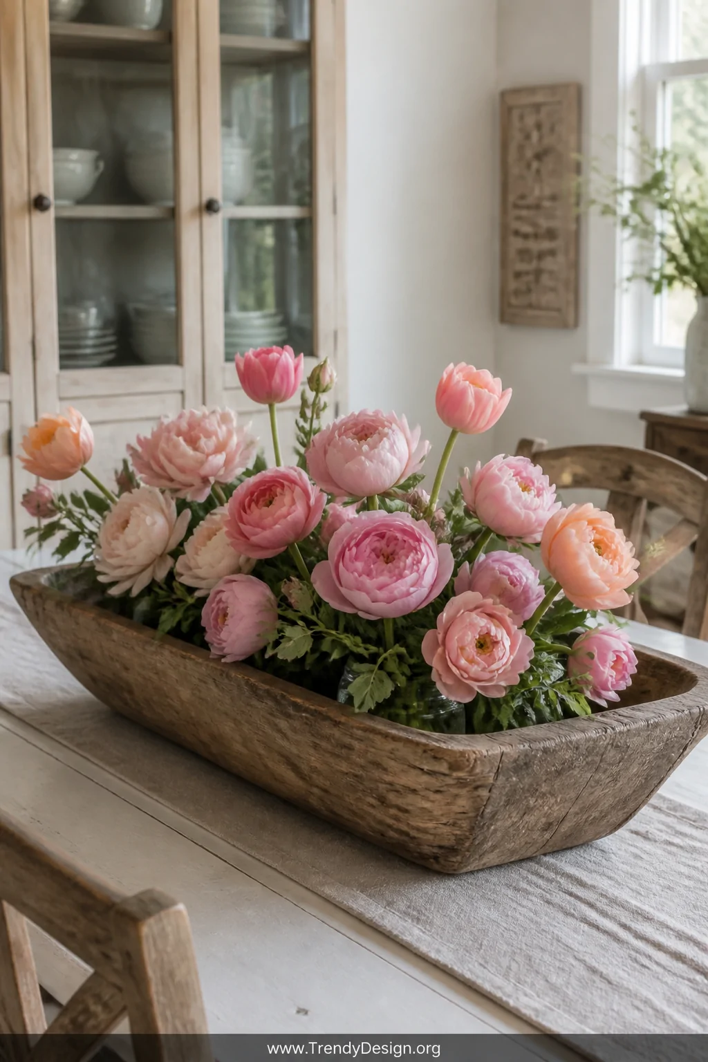 A medium shot of a weathered rustic wooden dough bowl on a white farmhouse dining table holding a lush arrangement of soft pi