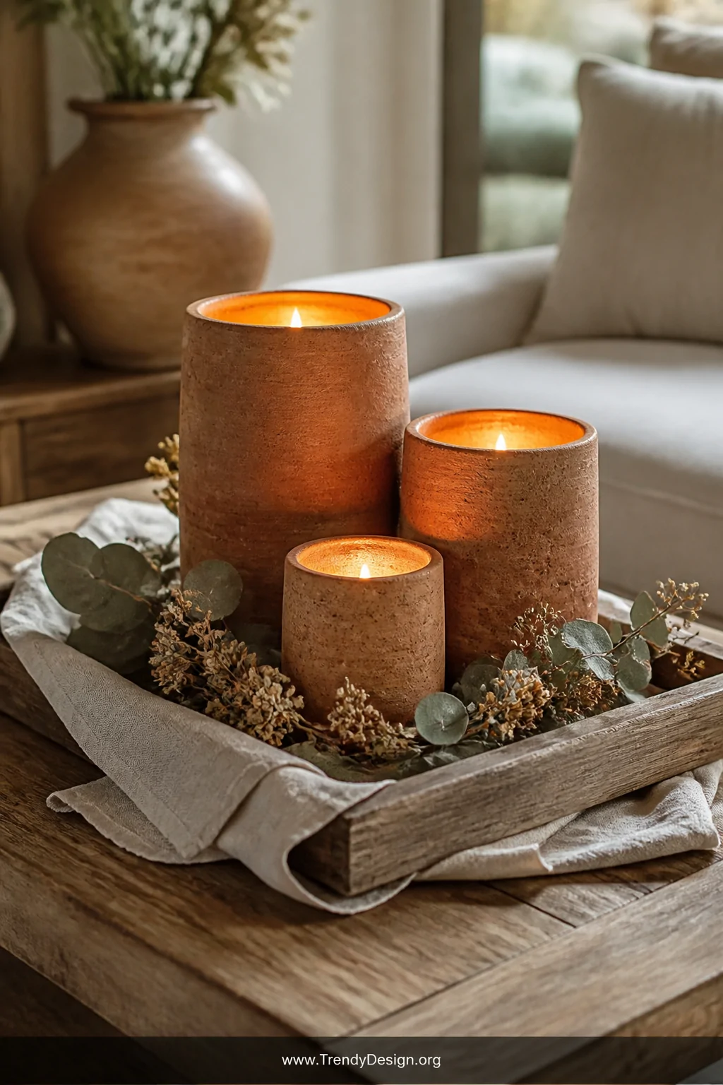 A closeup detail shot of a rustic wooden coffee table styled with three terracotta clay candle holders in varying heights glo
