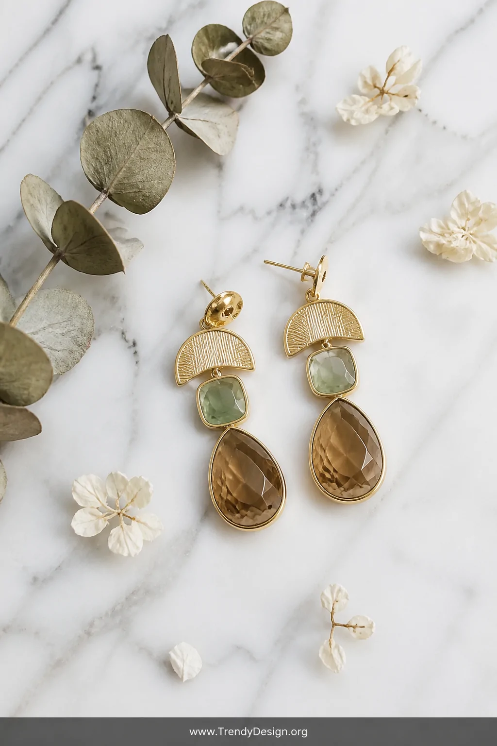 An overhead flat-lay shot of gold and gemstone earrings resting on a cool white marble surface with subtle grey veining, acco