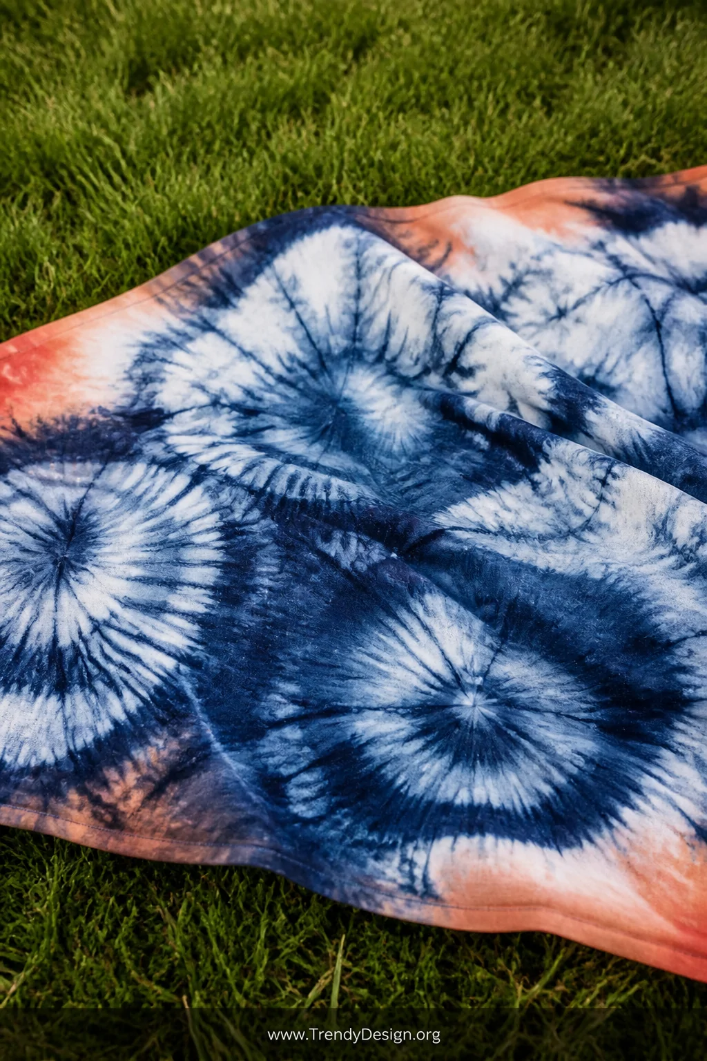 A close-up detail shot of an artisan hand-dyed indigo shibori picnic blanket laid on green grass, the organic swirling blue a