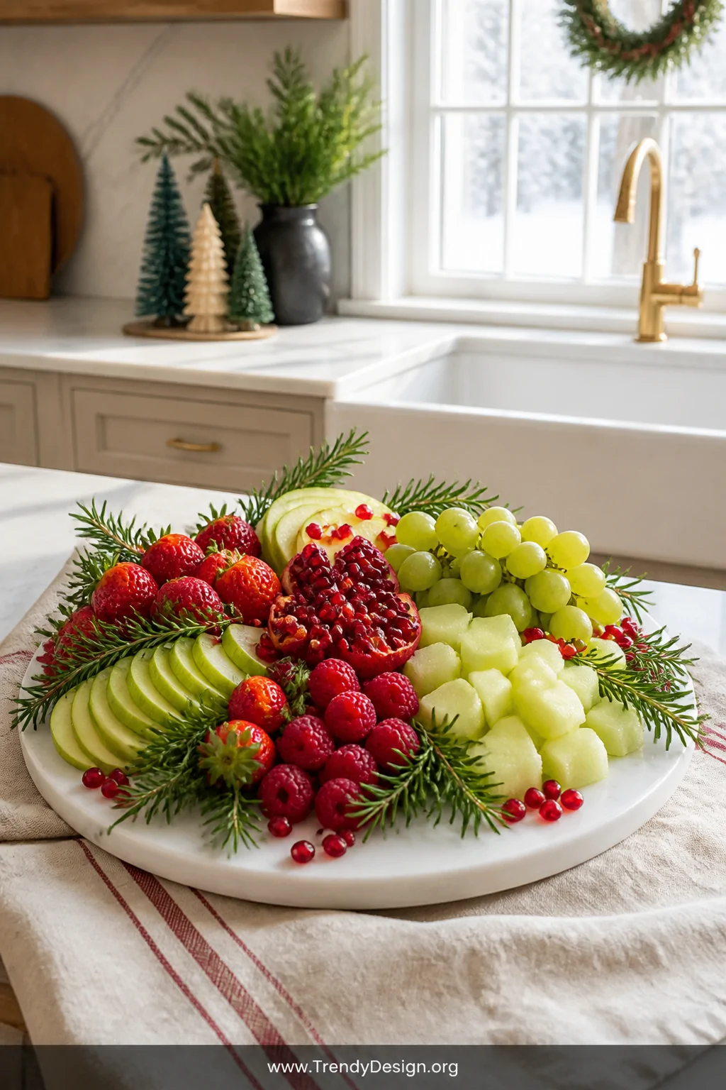 A medium shot of a red and green Christmas fruit platter displayed on a white marble board resting on a linen-draped kitchen 