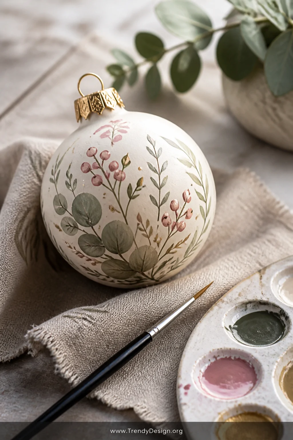 A close-up detail shot of a single cream ornament ball painted with delicate hand-illustrated botanicals including sage green