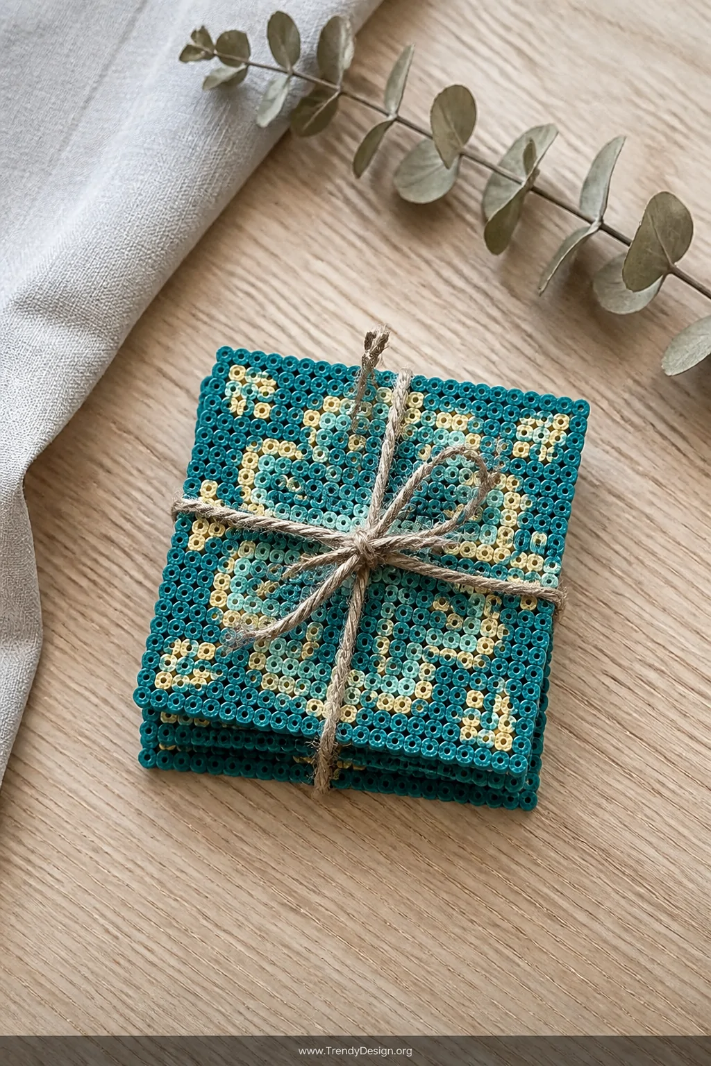 A medium overhead flat-lay shot of a set of four square Perler bead coasters in a cohesive teal and gold color scheme, stacke