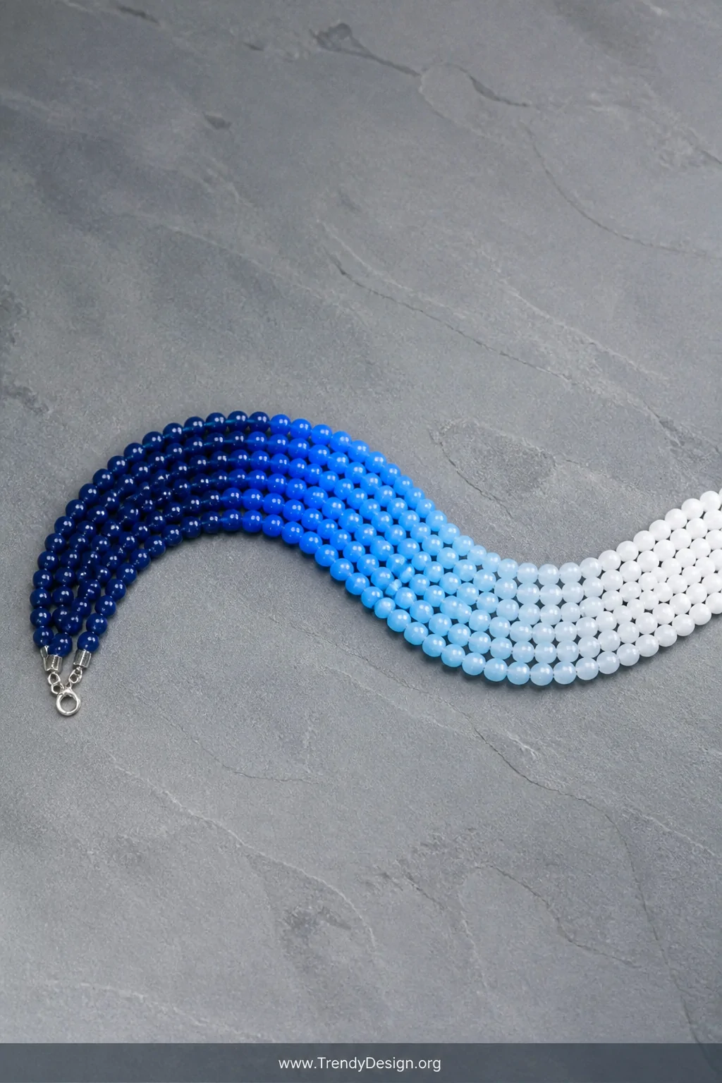 A wide flat-lay shot on a smooth slate gray surface featuring an ombre gradient pony bead necklace transitioning from deep na