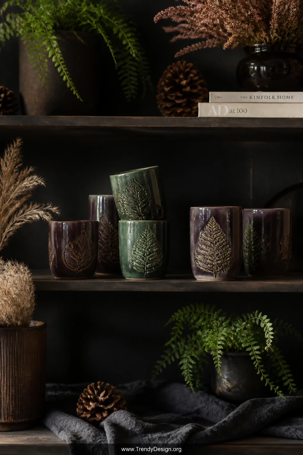 A wide shot of a dark and moody styled shelf scene featuring forest-green and deep plum glazed ceramic tumblers with pressed 