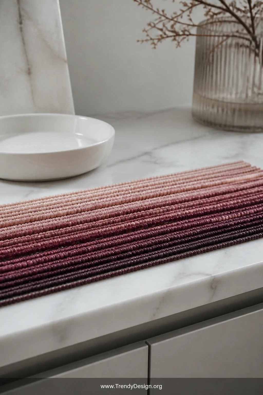 Close-up detail shot of a sleek marble vanity surface displaying a gradient arrangement of dusty rose to deep burgundy seed b
