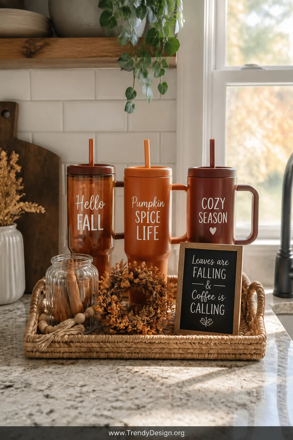A medium shot of a cozy kitchen counter tumbler station built on a woven rattan tray, featuring three fall tumblers in amber 