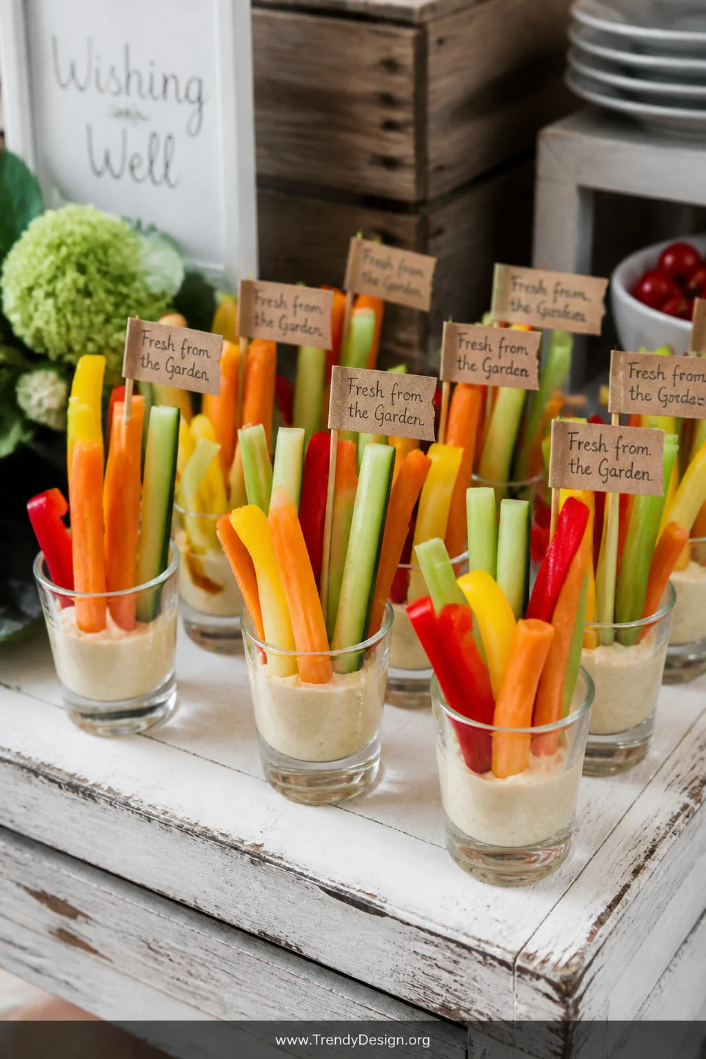 A close-up detail shot of a rustic snack table displaying small clear shot glasses filled with creamy beige hummus, with vibr