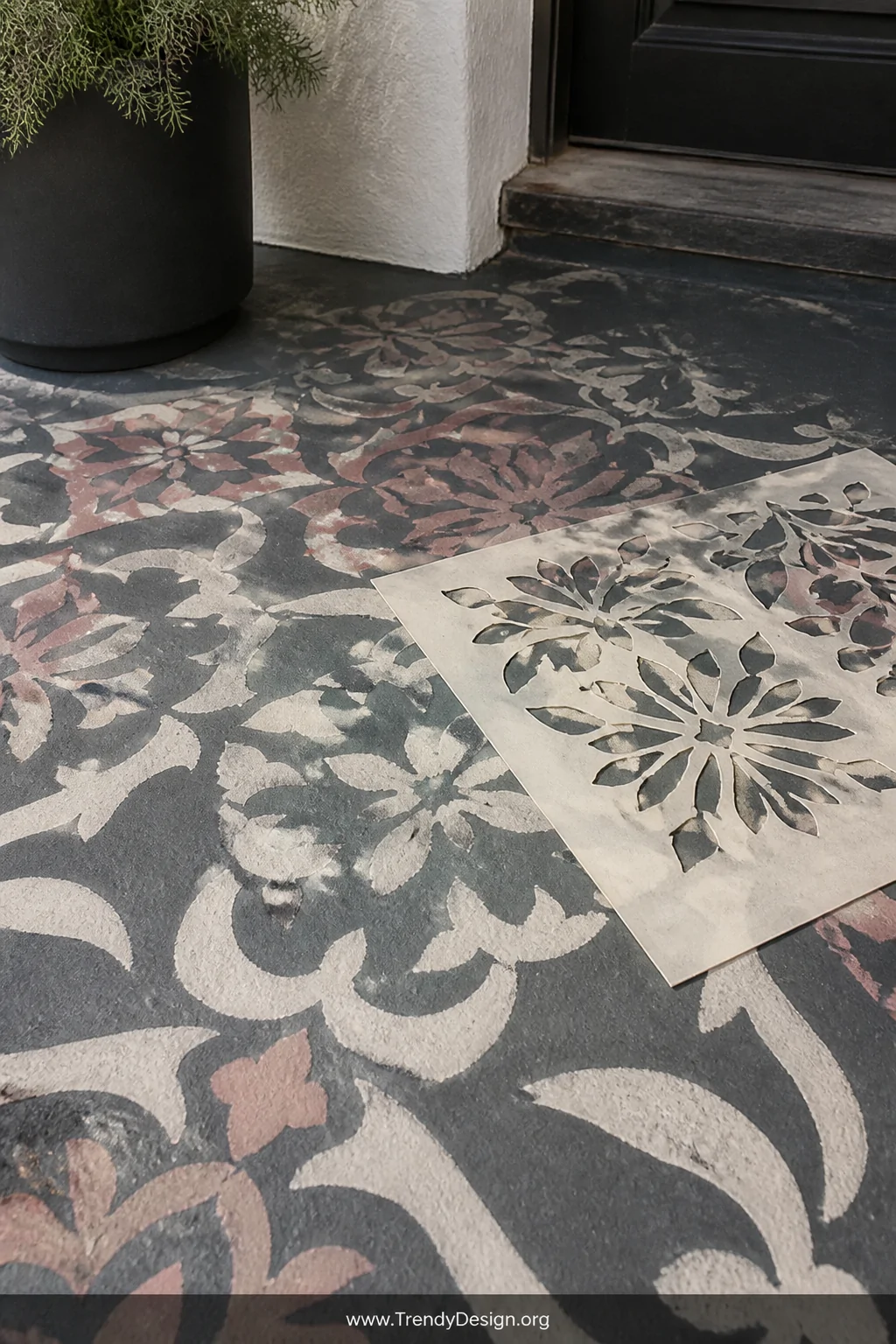 Close-up detail shot of a concrete porch floor mid-stencil reveal, showing an intricate Moroccan floral pattern in dusty rose