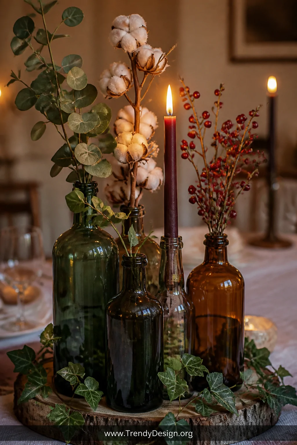 A close-up of a rustic dining table centerpiece featuring a tight cluster of 5 varying-height glass bottles holding eucalyptu