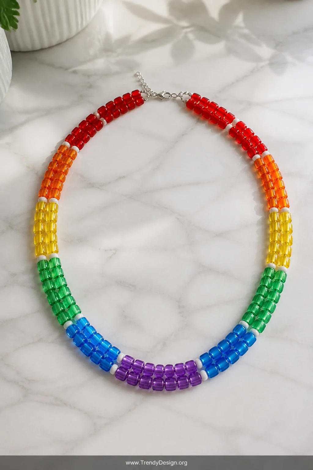 A wide flat-lay shot on a white marble surface displaying a finished rainbow stripe pony bead necklace with transparent red, 