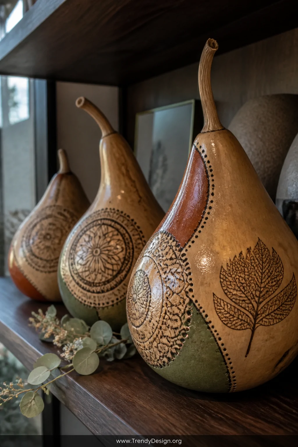A close-up detail shot of three dried gourds arranged on a dark walnut shelf displaying intricately burned mandala and lace-l