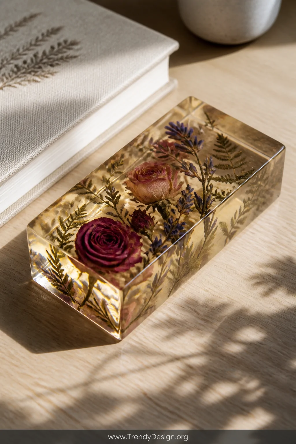 A close-up detail shot of a rectangular clear resin paperweight resting on a white oak desk, with pressed roses, lavender spr