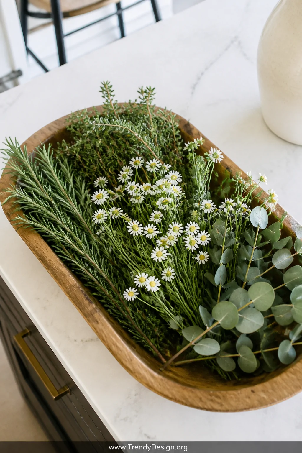 A medium overhead flat-lay shot of a wooden dough bowl on a kitchen island filled with an organic arrangement of fresh rosema