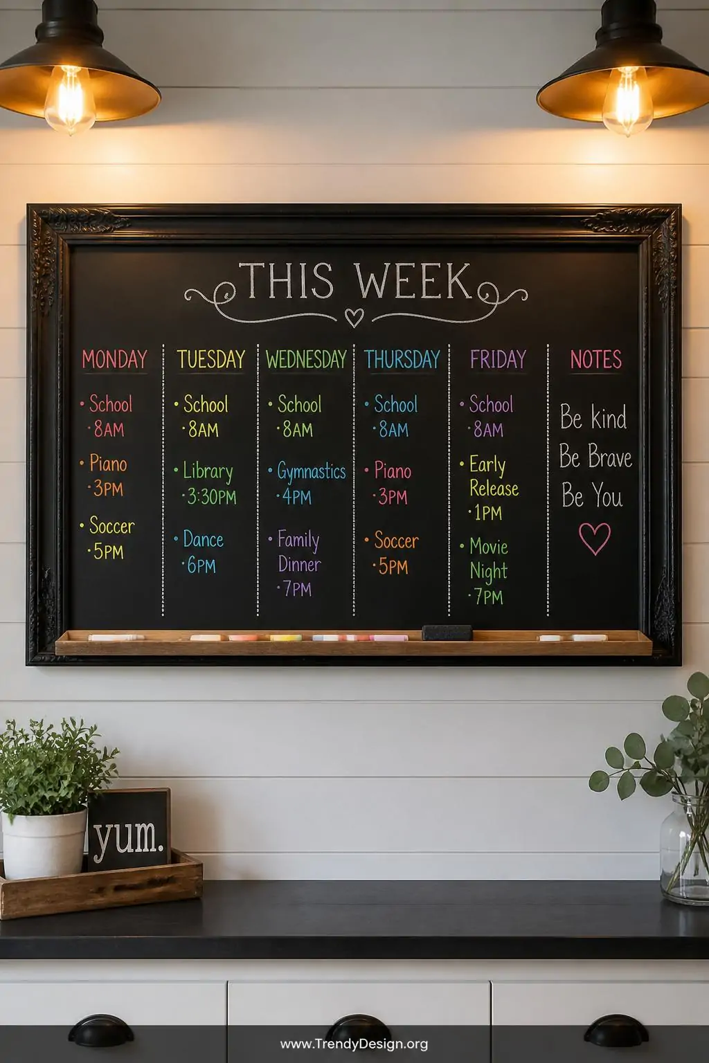 Large ornate black-framed chalkboard mounted on a white shiplap kitchen wall displaying colorful chalk marker weekly schedule divided into neat columns, warm pendant lighting casting a golden glow, medium shot showing the full frame with a slim wooden ledge below holding chalk and an eraser.